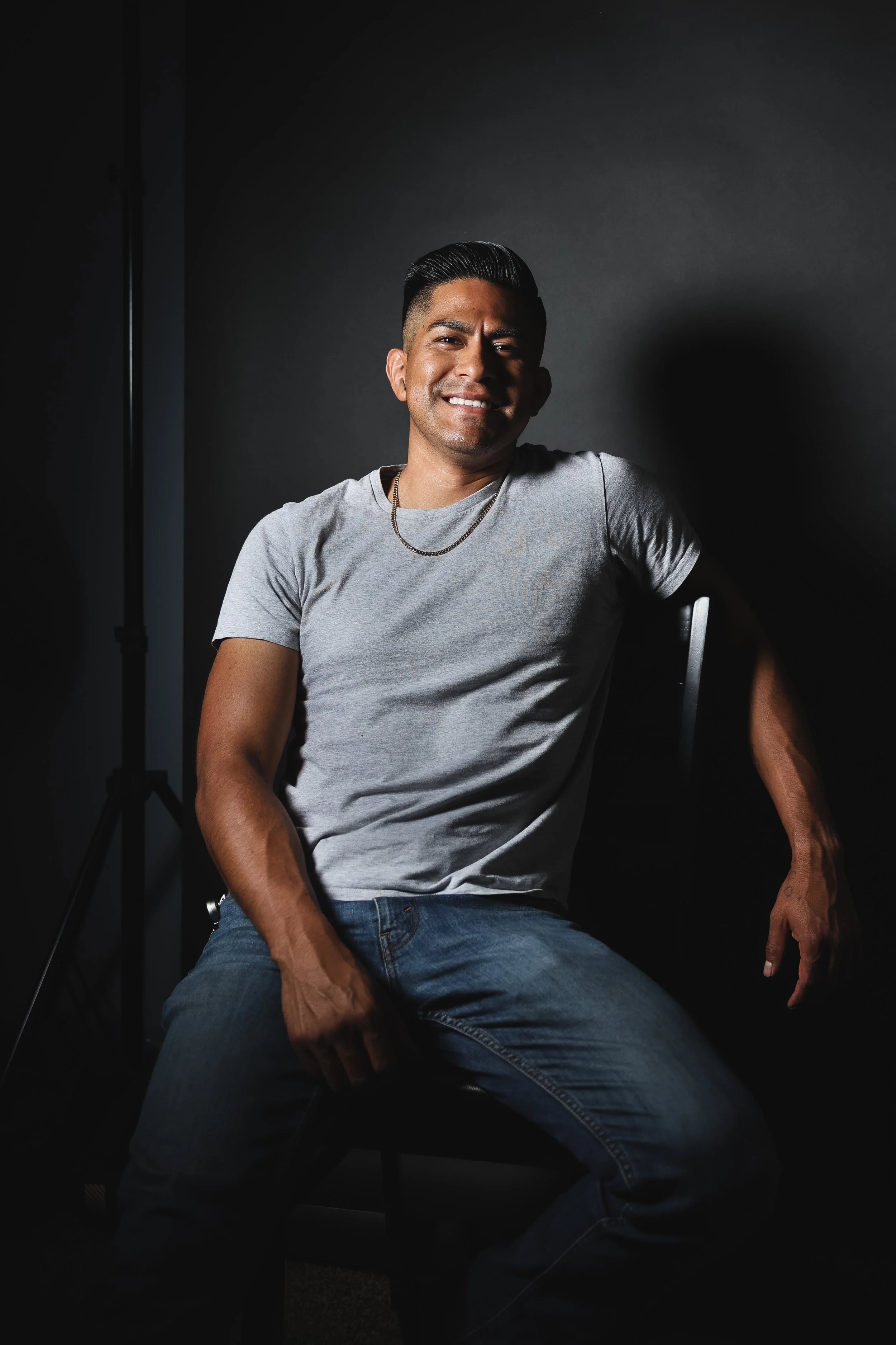 A young man with dark hair and tan skin, wearing a gray t-shirt and jeans, sitting on a chair against a dark background, smiling at the camera with a relaxed posture.