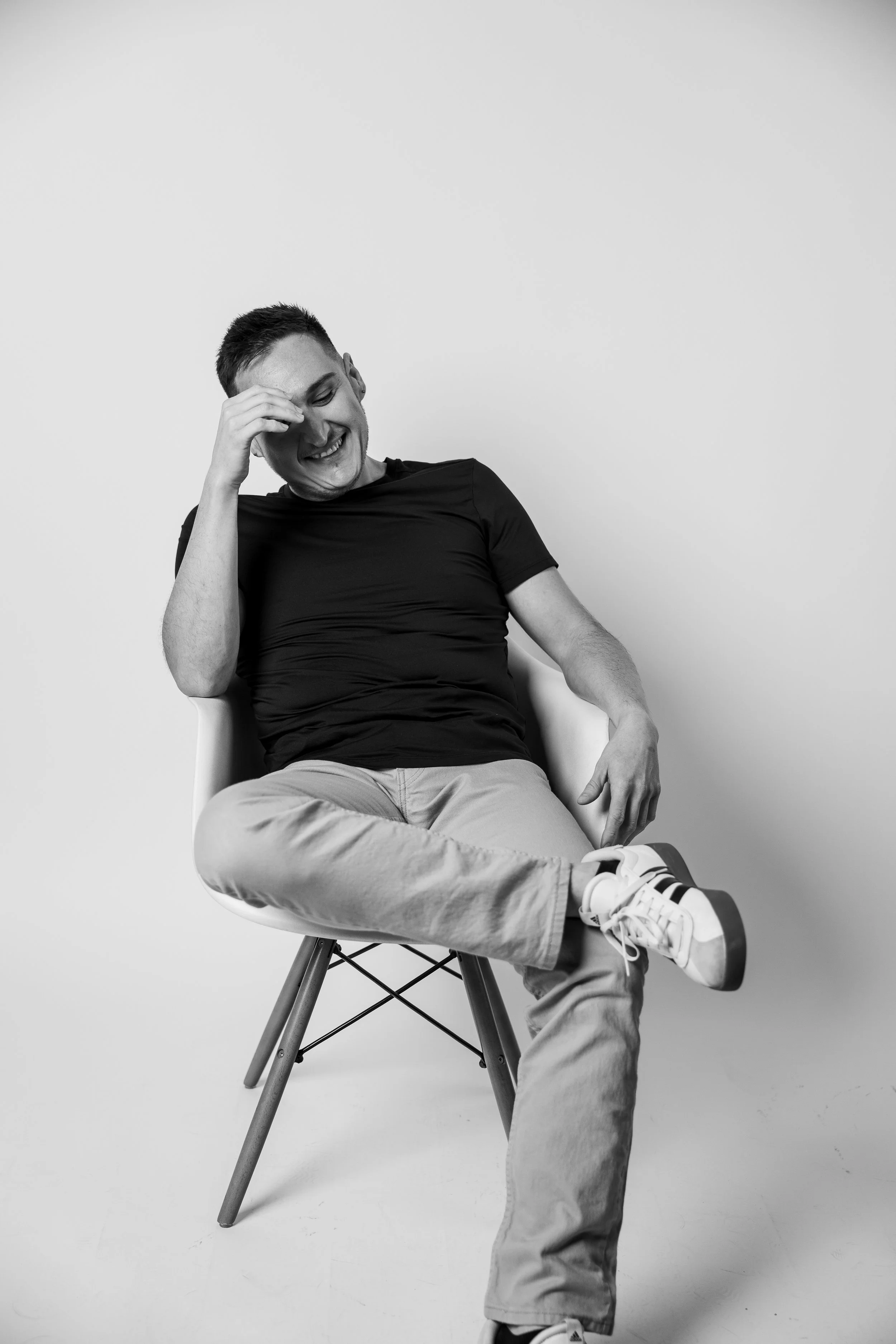 A young man sitting on a chair, smiling and laughing, wearing a black t-shirt, light-colored pants, and sneakers, against a plain white background in a black and white photo. Riverside Portraits