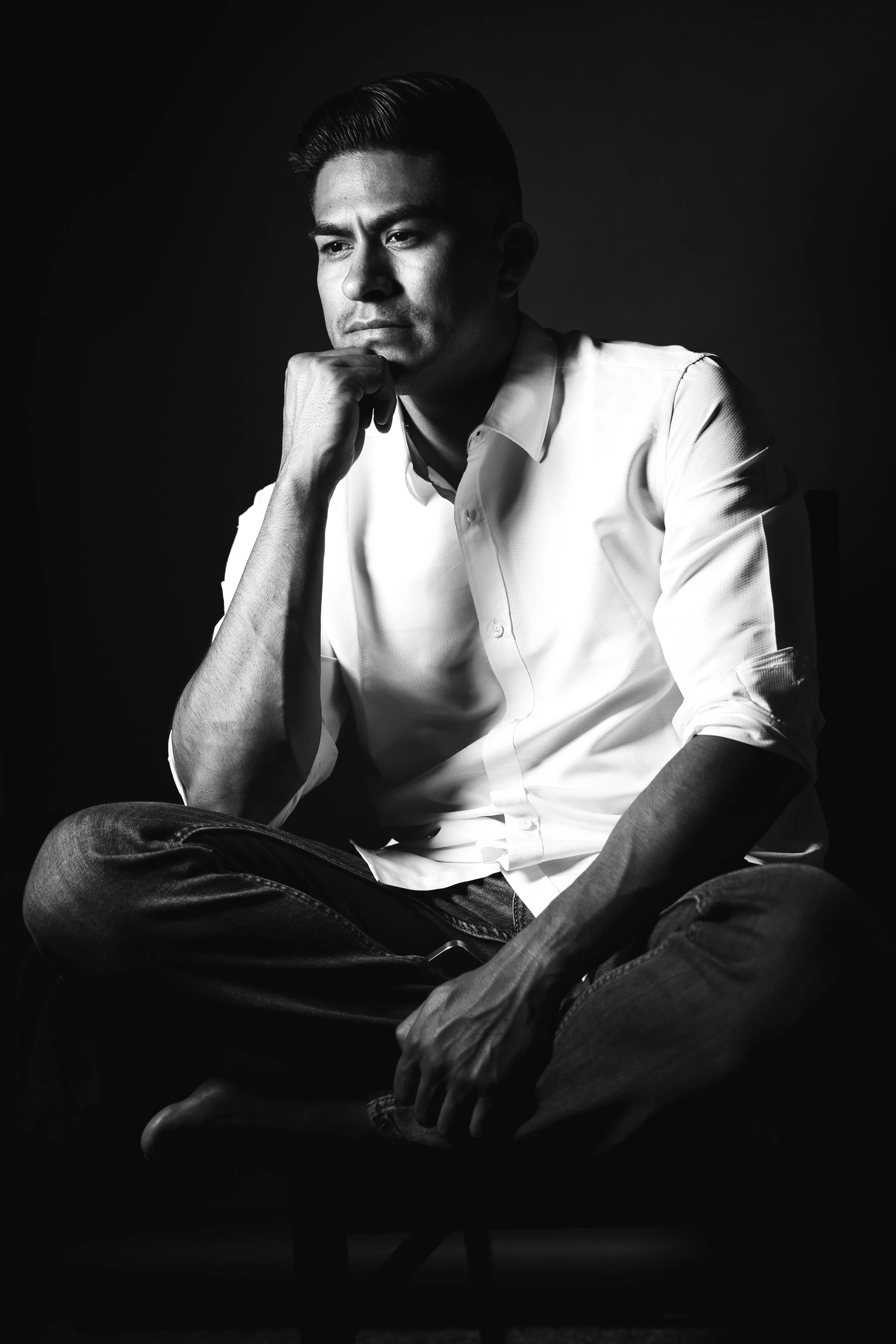 Black and white portrait of a man sitting cross-legged, wearing a white shirt, with his hand resting on his chin, looking thoughtfully into the distance. Riverside Portraits