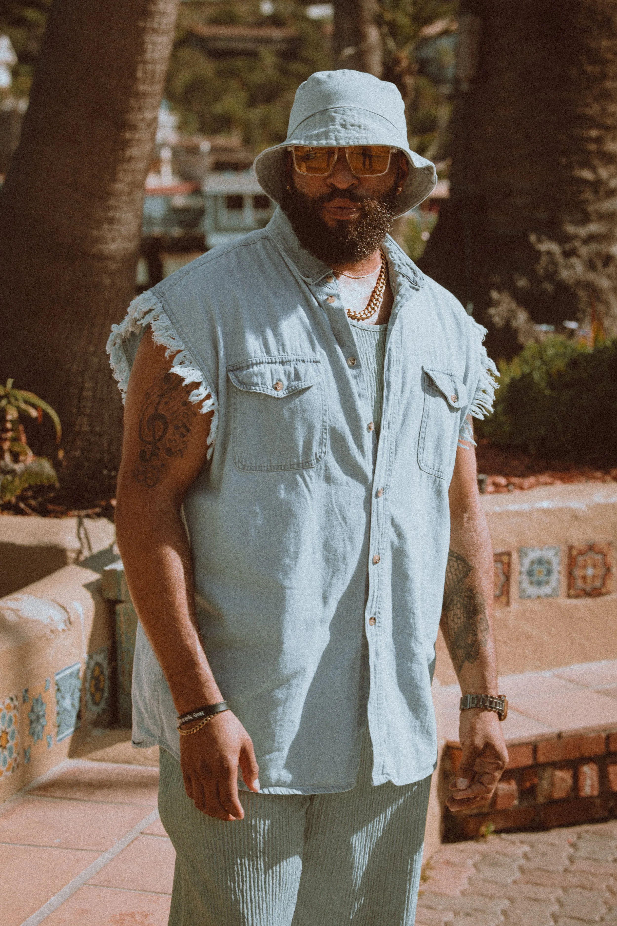 A man with a beard wearing a light denim sleeveless shirt, a bucket hat, sunglasses, and gold chains, standing outdoors near palm trees.