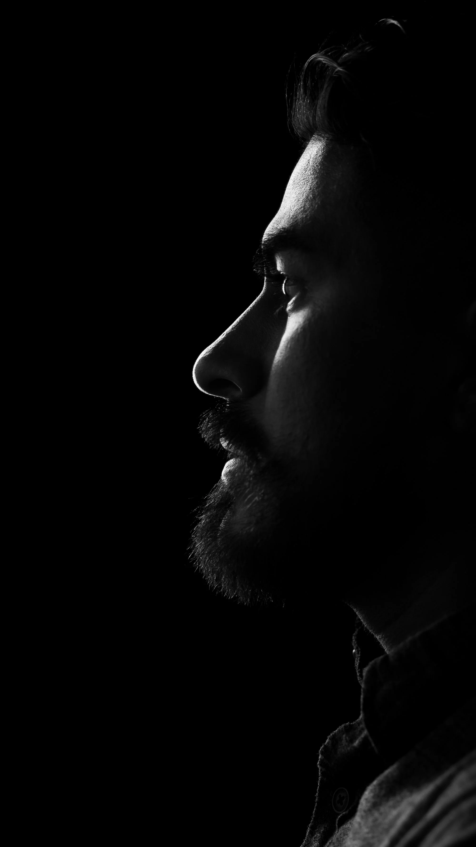Black-and-white side profile of a man's face, with dramatic lighting highlighting his nose, lips, eye, and beard against a dark background. Riverside Portraits