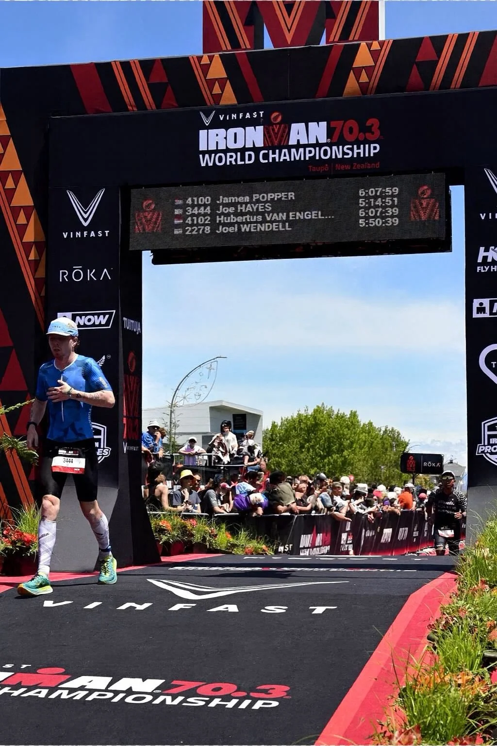 A male athlete crossing the finish line at the Ironman 70.3 World Championship in Taupo, New Zealand, with a digital scoreboard displaying his and other competitors' names and times, and a crowd of spectators behind the barrier watching the race.