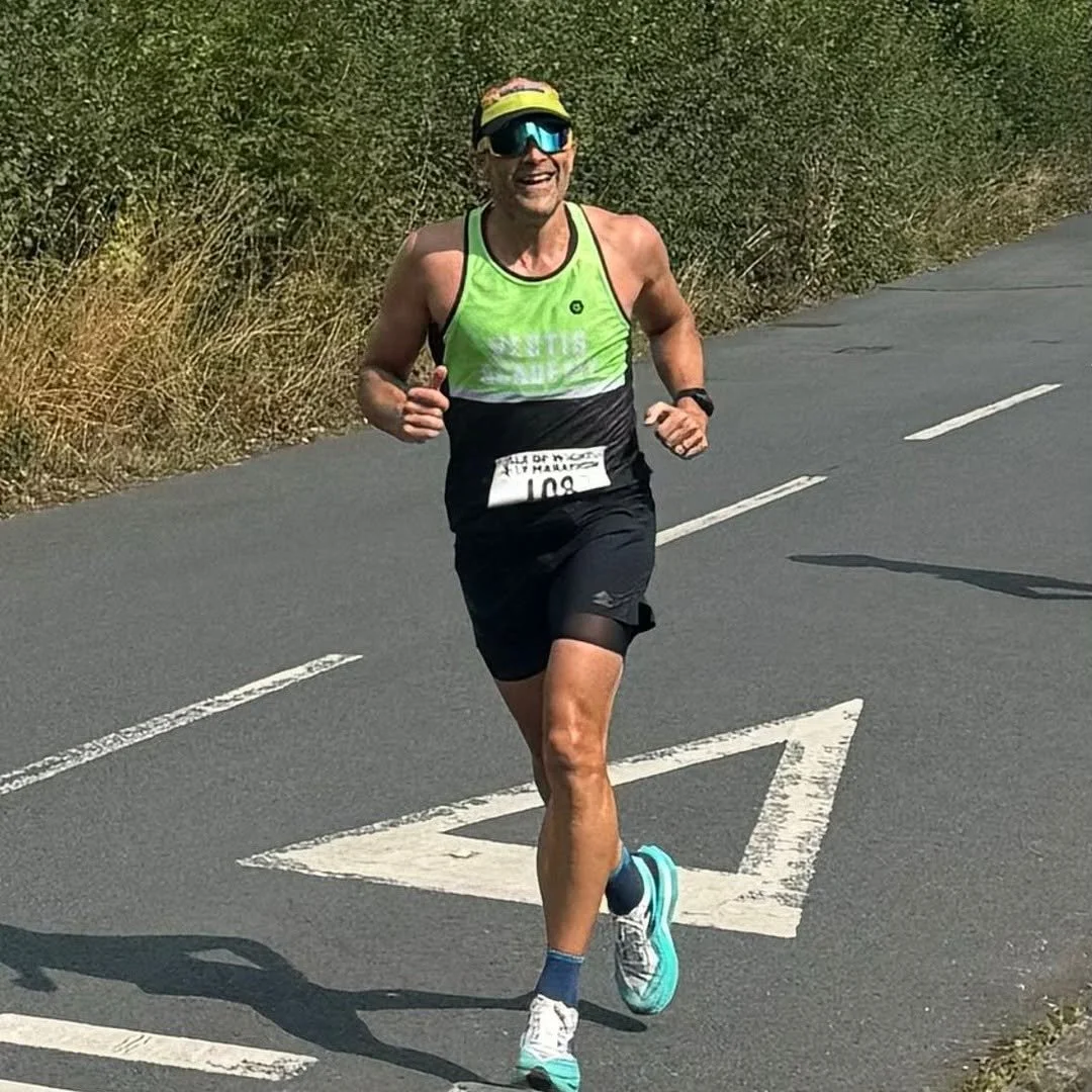 A person running on a paved road with dry grass and bushes on the side, wearing a bright green and black athletic shirt, black shorts, blue running shoes, a yellow visor, sunglasses, and a race bib.