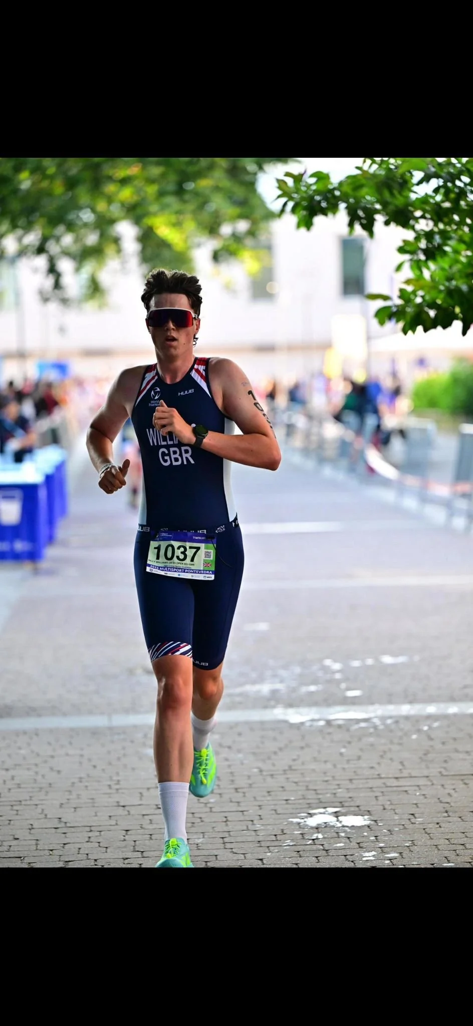 Female athlete in medal, sunglasses, black hair, running in a race, wearing blue sports outfit with GB and number 1037, white socks, neon green shoes, race bib, outdoors with trees and blurred spectators in background.