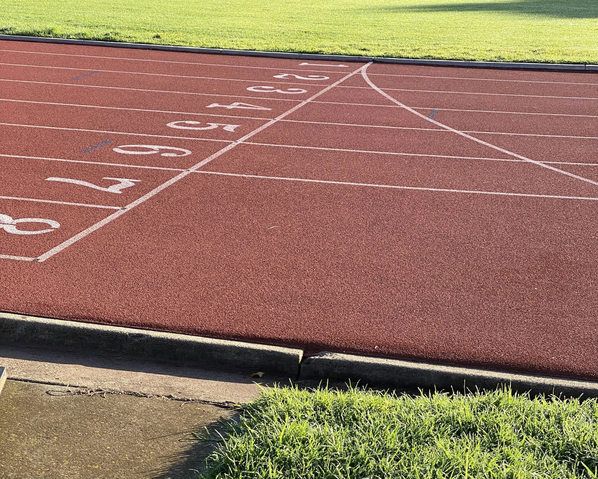 A section of a red running track with numbered lanes from 0 to 5, and a curved white line marking the starting point. Green grass is visible at the bottom of the image.