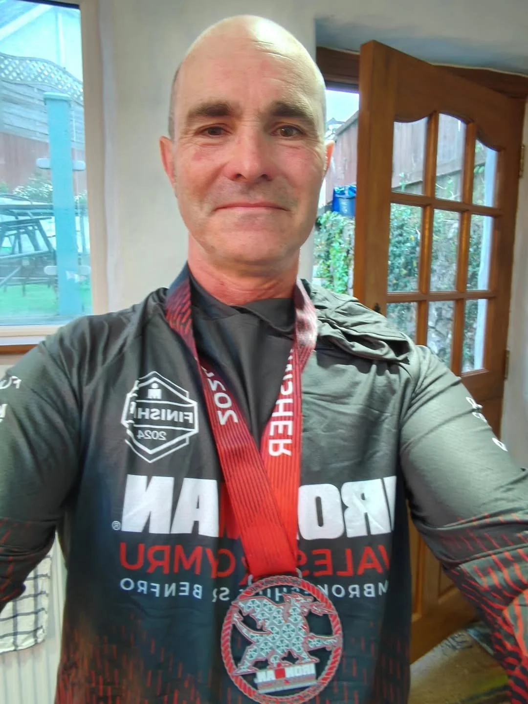 Man taking a selfie indoors, wearing a black and red sports t-shirt with a medal around his neck, standing near a wooden door with glass panes and a window in the background.