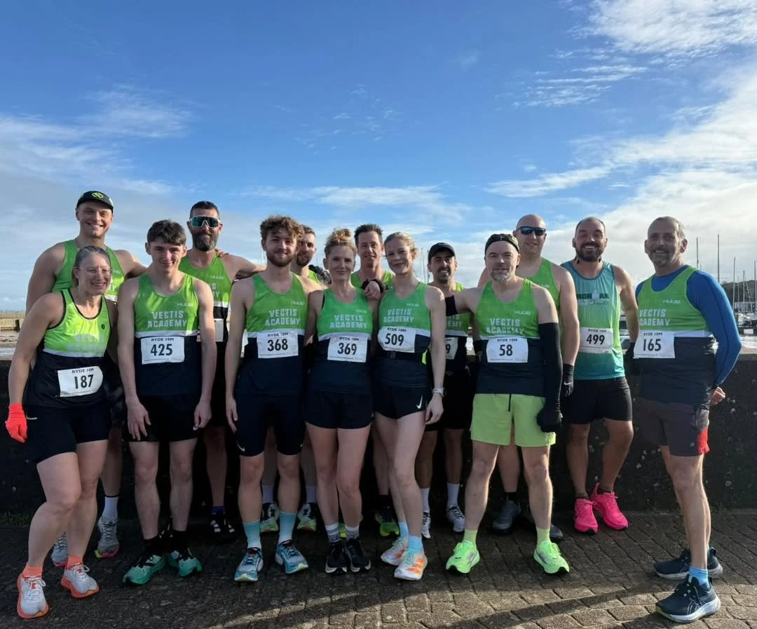 Group of runners in matching green and black athletic gear posing outdoors before a race, with some wearing bibs and a sky with clouds in the background.