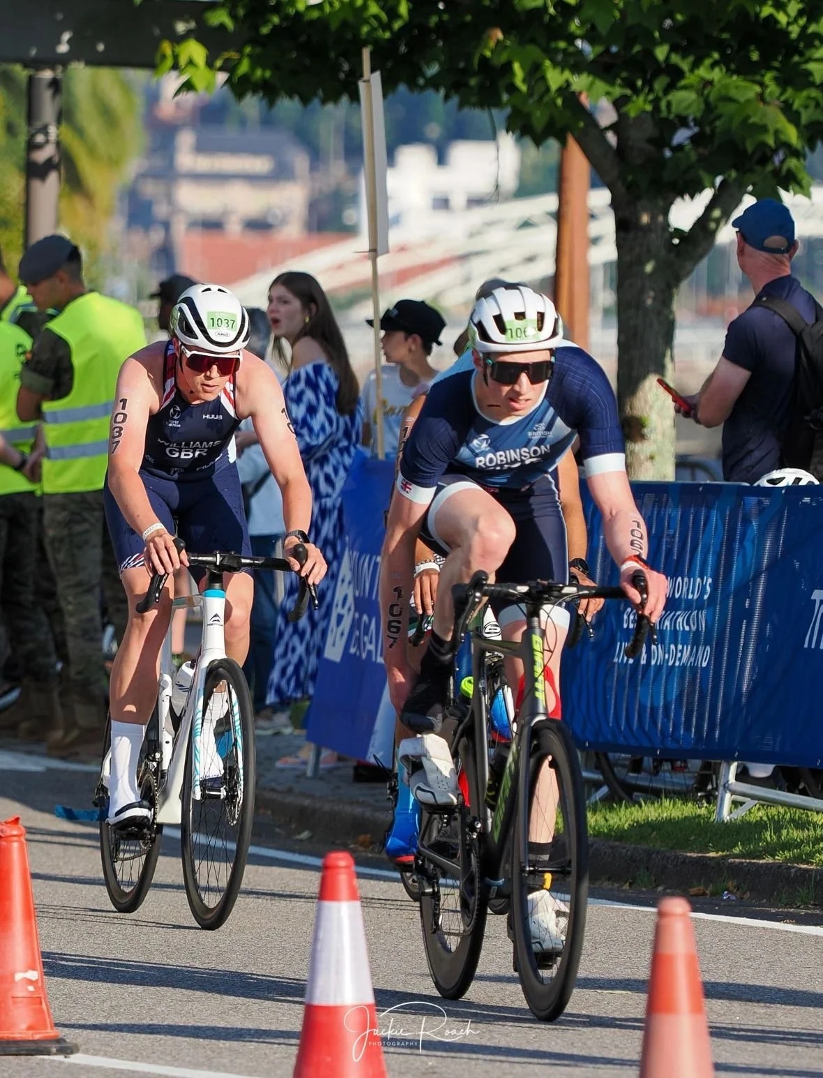Two cyclists in a race, wearing helmets and sunglasses, riding on the road between orange traffic cones, with onlookers and event staff in the background.