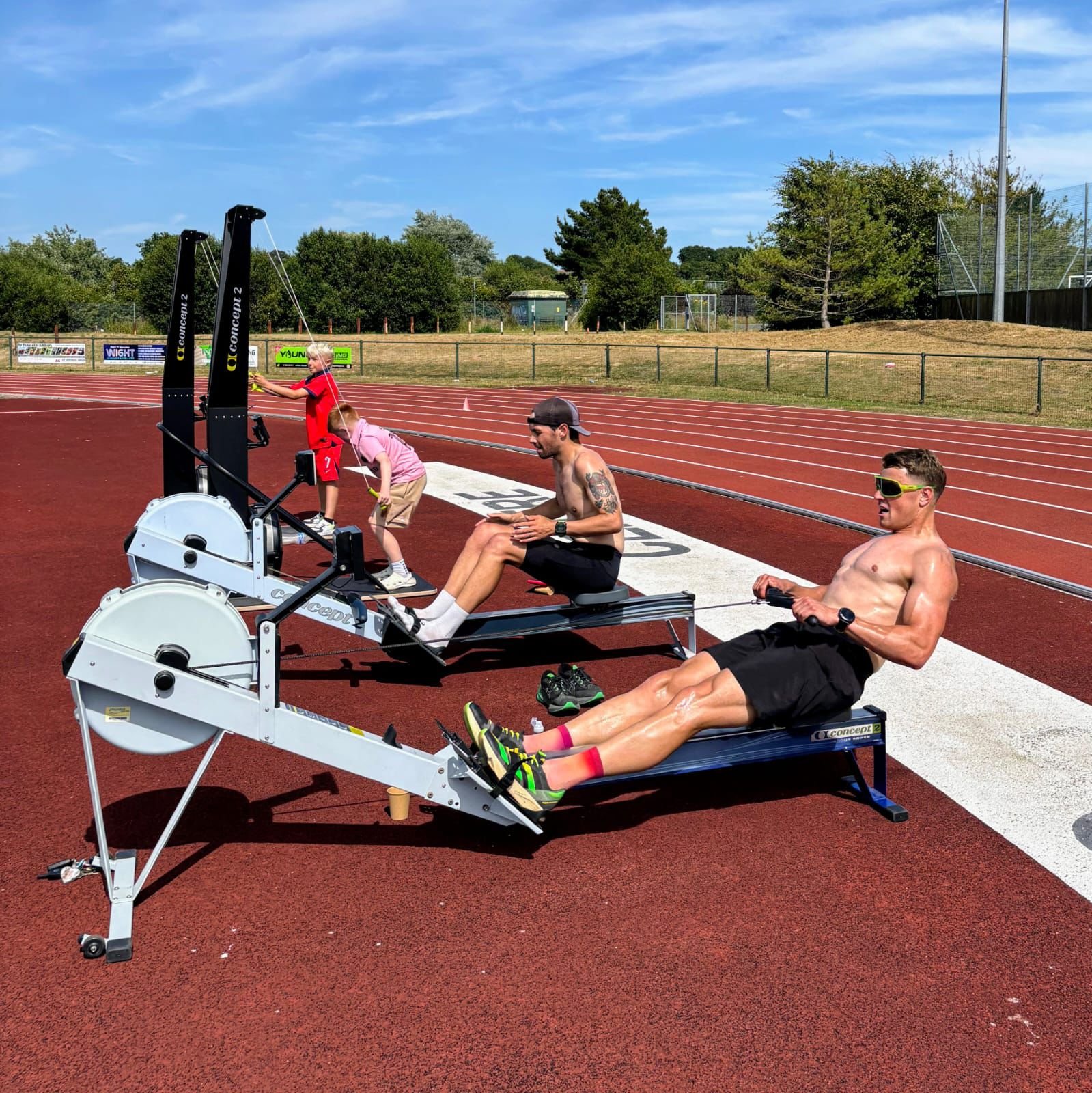 Two men and two children using rowing machines on a red outdoor track on a sunny day, with green trees and a blue sky in the background.