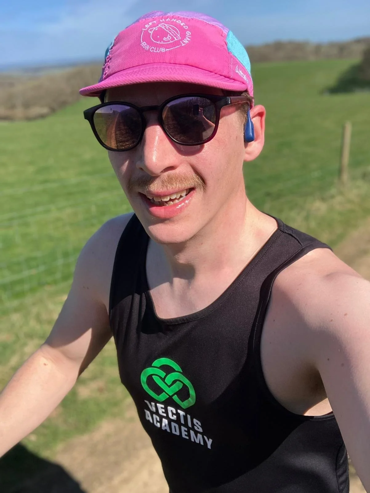 A man wearing sunglasses, a pink cap, and a black tank top with a green and white logo that says 'Vectis Academy' takes a selfie outdoors on a sunny day. He is smiling, and the background features green grass and rolling hills.