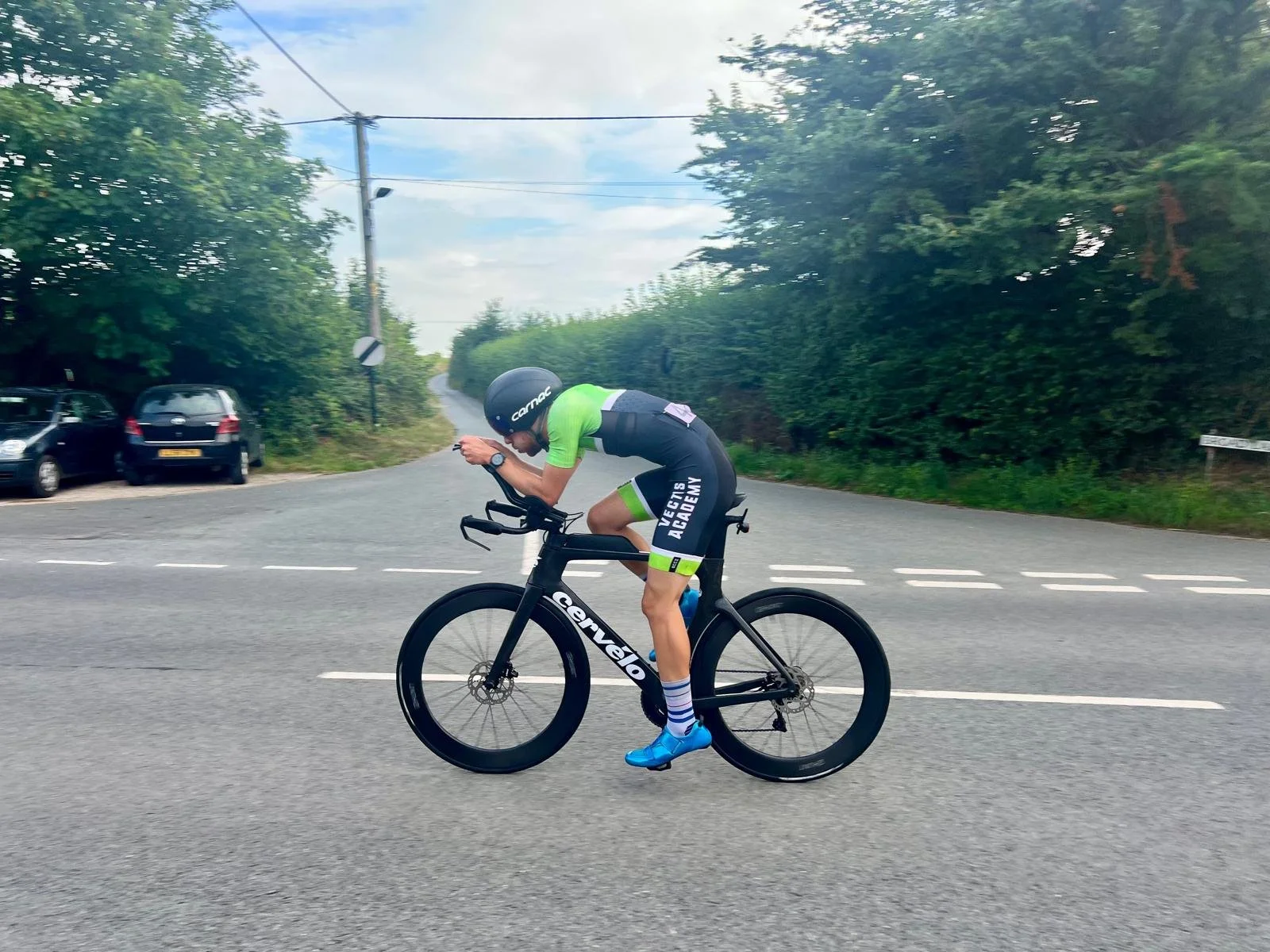 A cyclist wearing a black helmet, black and green cycling suit, and blue shoes riding a black Cervélo bike on a paved road with greenery on both sides.