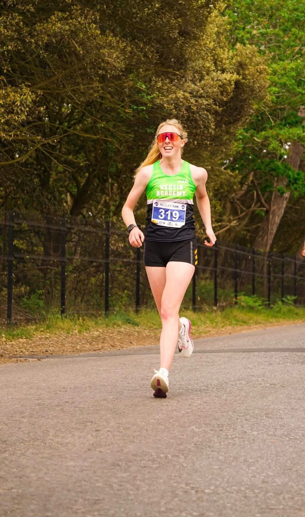 A woman running in a marathon, wearing a green tank top with 'VECTIS ACADEMY' printed on it, black shorts, white running shoes, red sunglasses, and a race bib number 319.