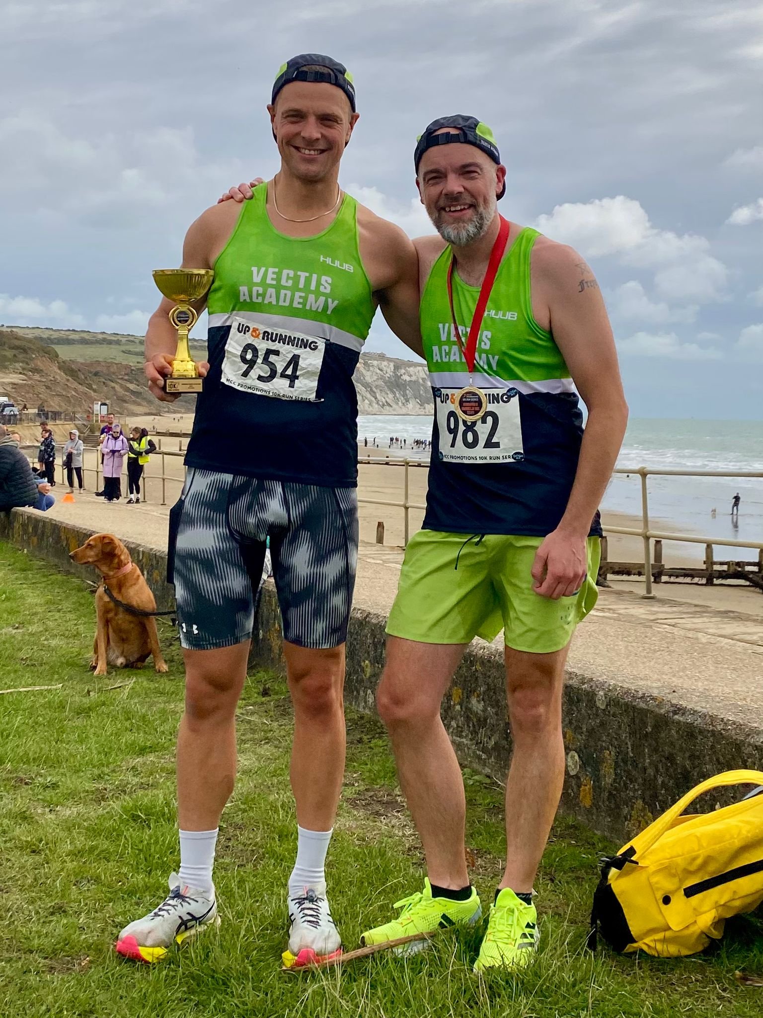 Two men in green and black running outfits celebrating at a beach, with one holding a trophy, and the other wearing a medal around his neck, after a race. A dog is sitting on the grass nearby, and there are spectators and the ocean in the background.