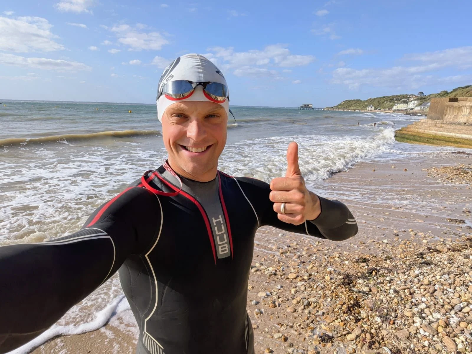 A smiling man in a wetsuit taking a selfie on a rocky beach, wearing a swim cap and goggles, with the ocean, waves, and a distant pier visible in the background.