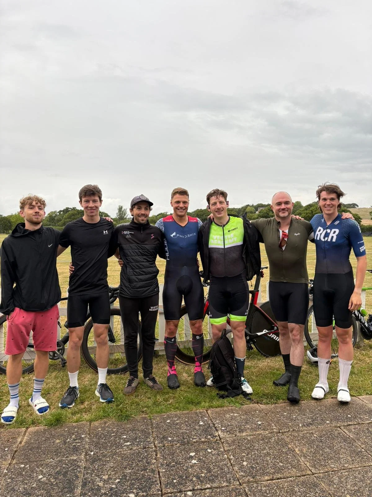 Group of seven men standing outdoors on a cloudy day, some in cycling gear with bicycles behind them, smiling and with arms around each other.