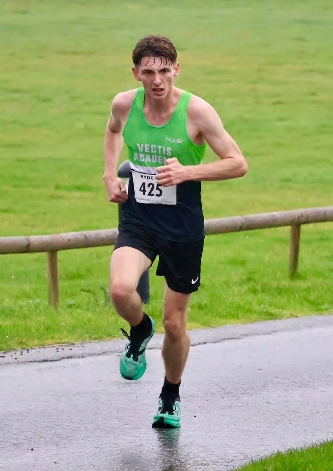 Male runner wearing a green tank top and black shorts running on a wet path during a race.
