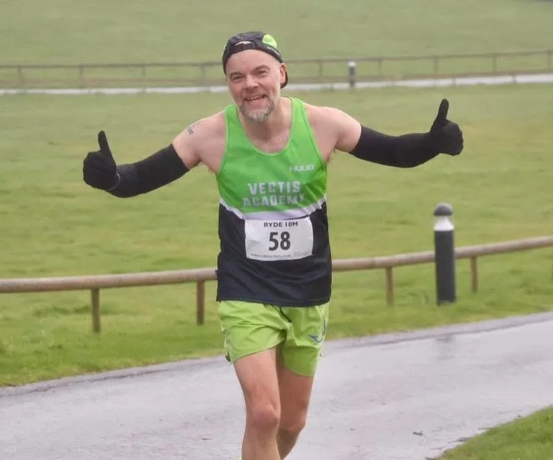 A man wearing athletic gear, including a green tank top, lime green shorts, black gloves, and a backwards cap, running outdoors on a path in a park, giving a thumbs-up with both hands, smiling, with a race bib numbered 58.