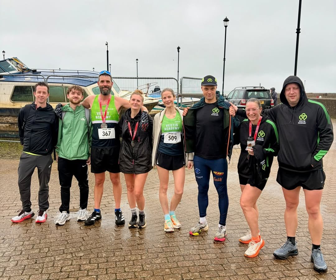 A group of nine runners poses outdoors on a damp, paved surface near boats and parked cars on a cloudy day. They are dressed in athletic gear, some with medals, and stand close together, some with arms around each other.