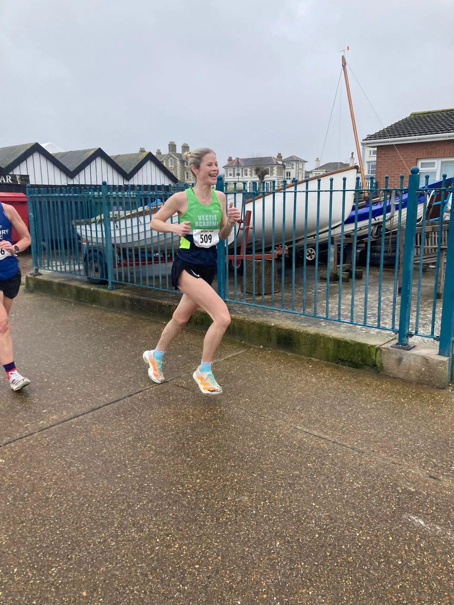 A woman running in a race, wearing a green and black tank top with the words 'VECTIS ACADEMY' and the number 509 on her bib. She is smiling and appears to be enjoying the race, with a cloudy sky and boats on land behind her.
