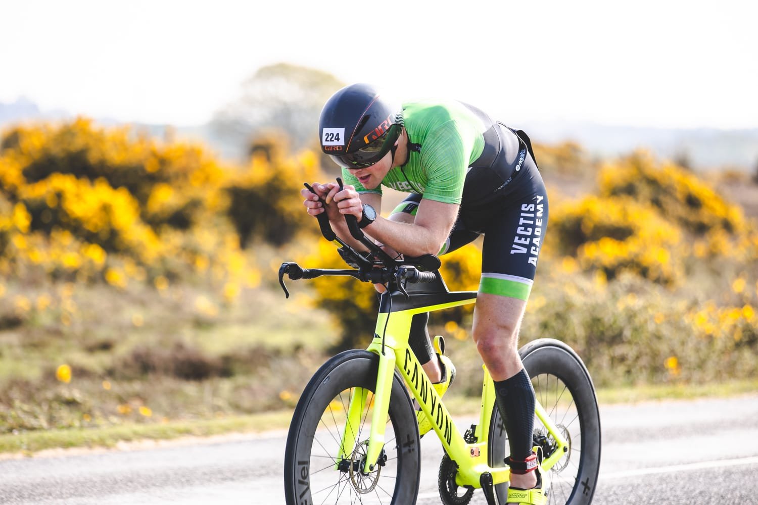 Cyclist wearing a green and black uniform, helmet, and bike, leaning forward and using a device on the handlebars while riding on a road with yellow flowers in the background.