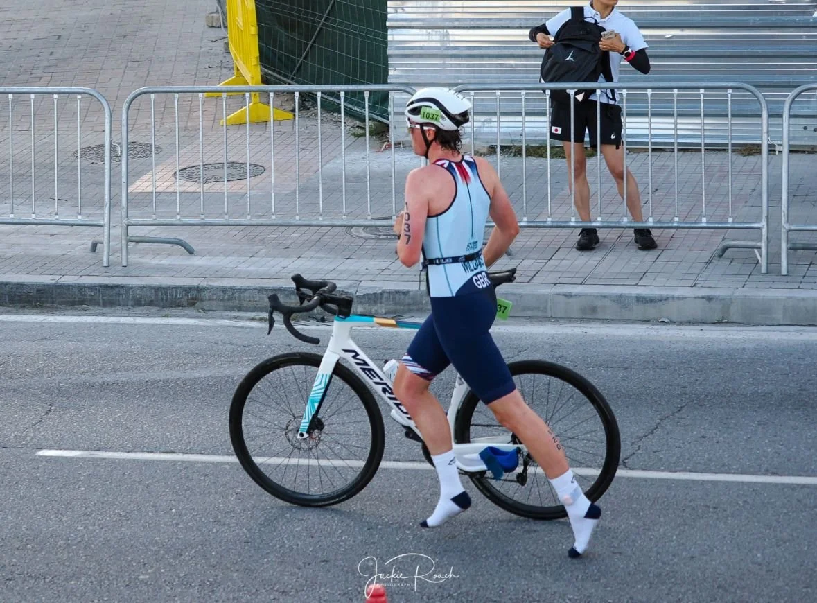 A female athlete in a blue and white triathlon suit, wearing a white helmet, standing next to her white and blue racing bike on a city street. Behind her, a young man with a backpack is standing behind metal barriers, looking at his phone.
