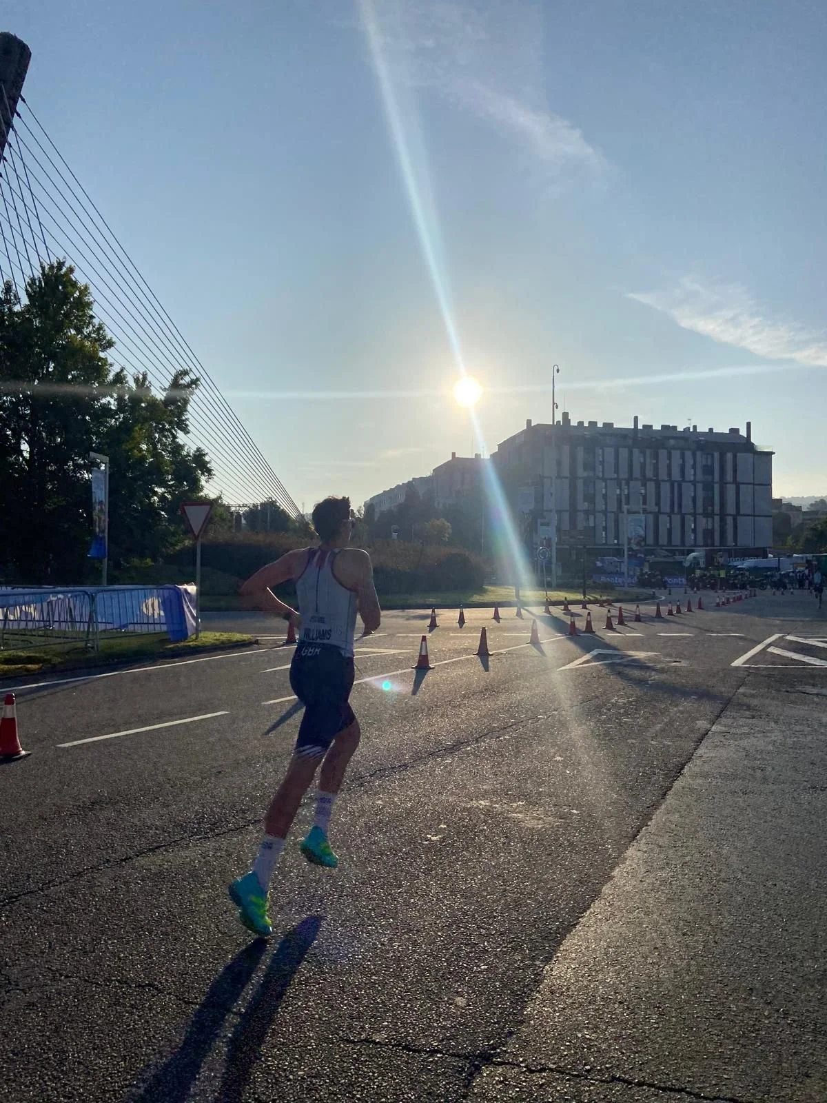A male runner running on a road during a sunny day with the sun low in the sky, casting long shadows. He is wearing a white tank top and dark shorts, with bright running shoes. There are traffic cones and barriers along the route, and a large buildin