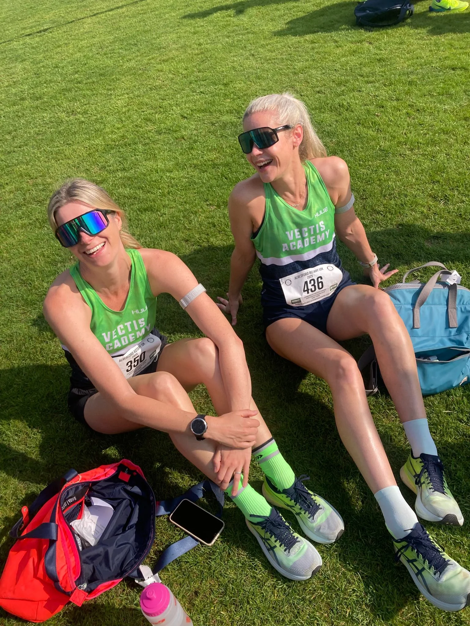Two women sitting on grass after running, wearing green and black athletic uniforms with bib numbers, sunglasses, and running shoes, smiling and talking.