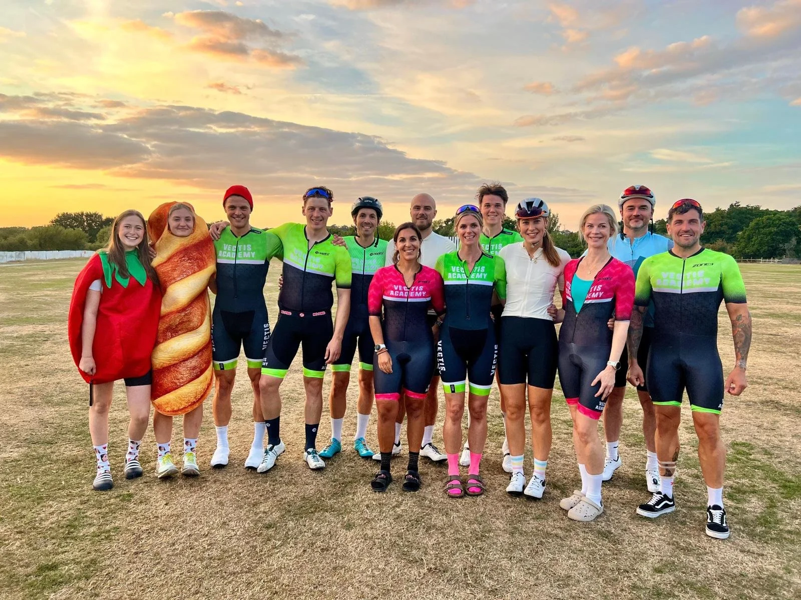 Group of cyclists and two people dressed as a tomato and a chili pepper standing on a grassy field during sunset.