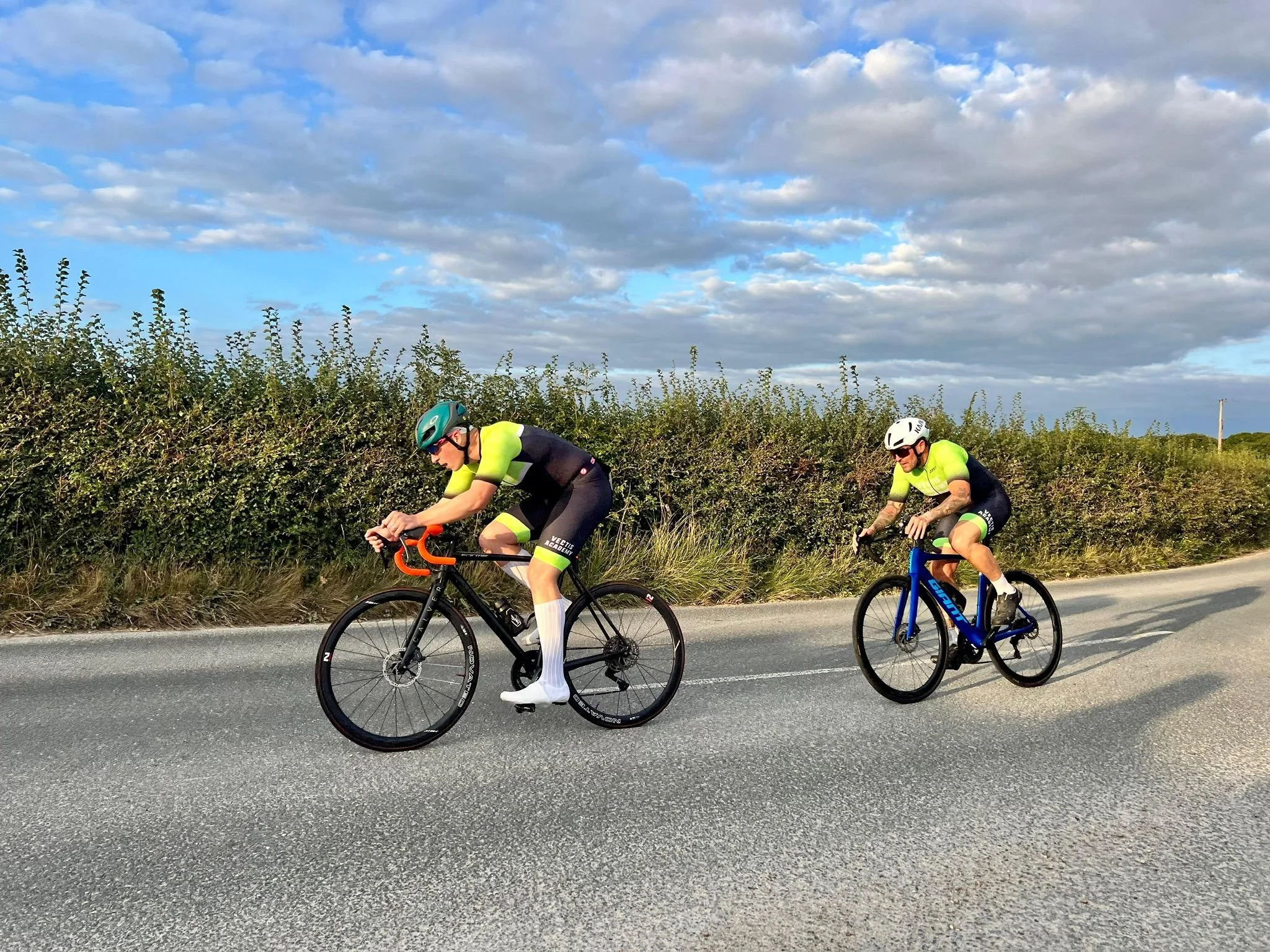 Two cyclists riding on a paved road during the daytime with a backdrop of green bushes and a blue sky with clouds.