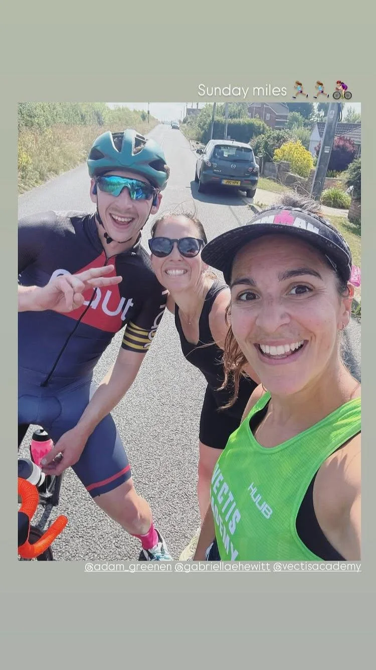 Three smiling women taking a selfie on a sunny day during a cycling activity, standing on a residential street with cars parked along the side.