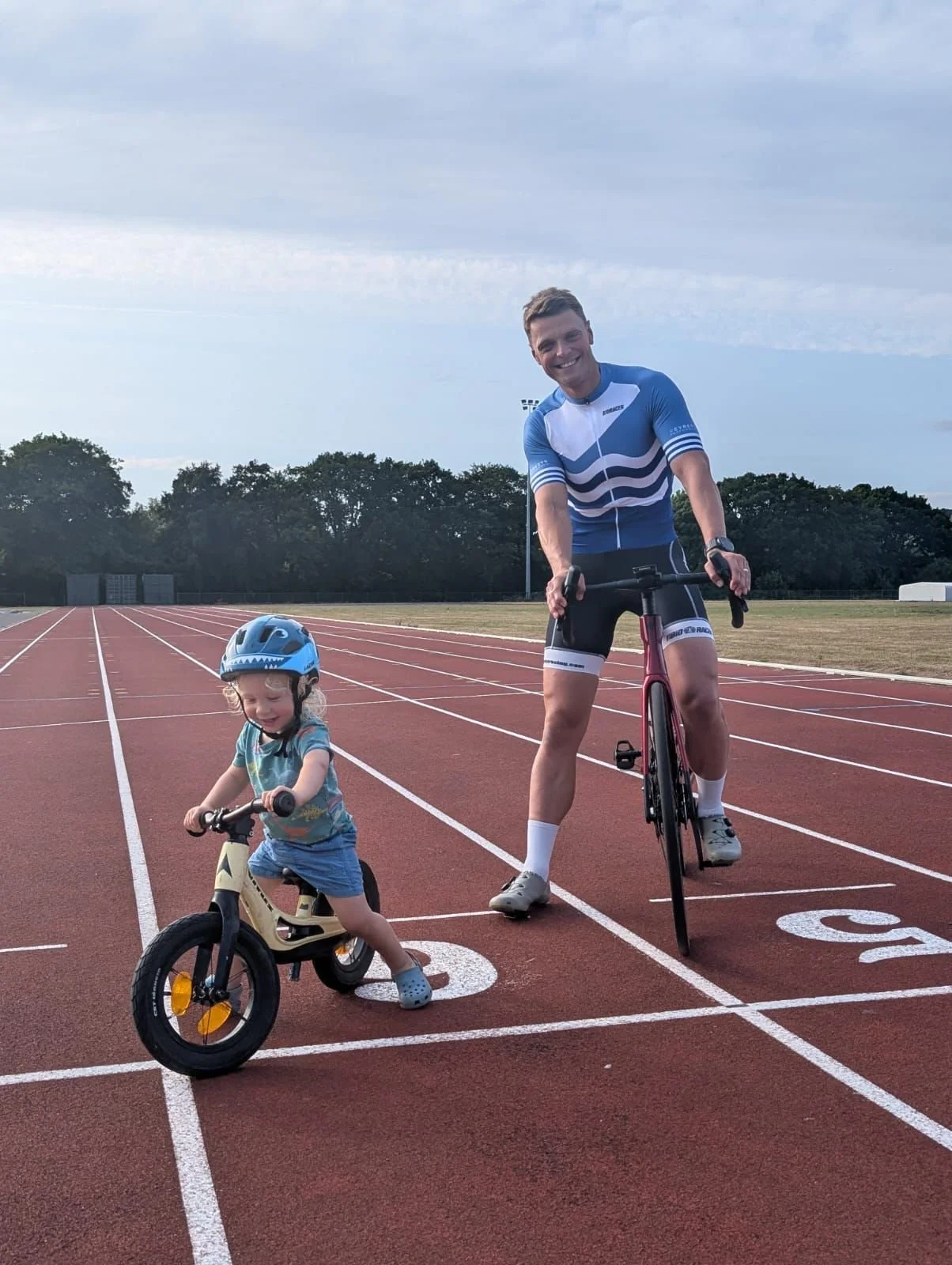 A young boy and an adult man riding bikes on a running track outdoors. The boy is on a small balance bike, wearing a helmet and a colorful shirt, smiling. The man is standing on an inline skate, smiling, in athletic clothing.
