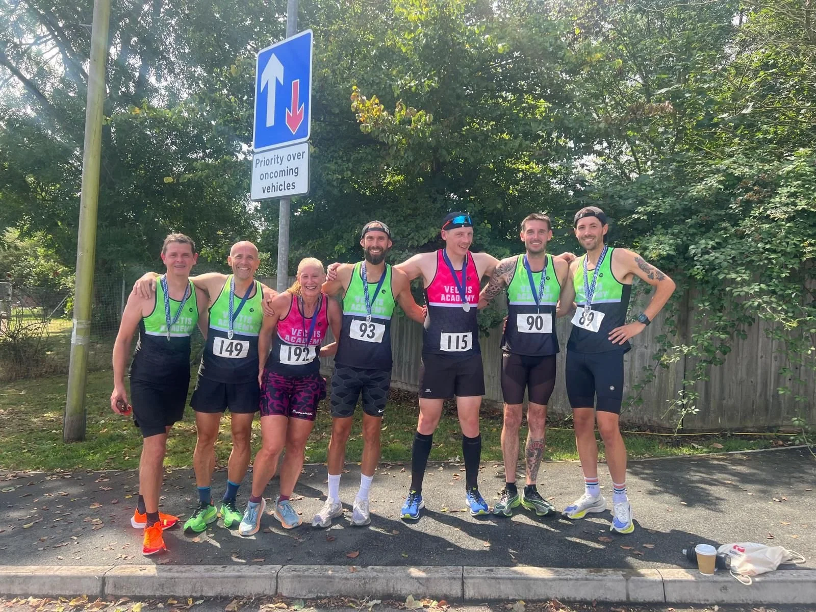 Group of six marathon runners standing on street with race medals, wearing athletic gear, some with numbers, smiling with arms around each other. Behind, trees and a blue trench sign indicating priority over oncoming vehicles.