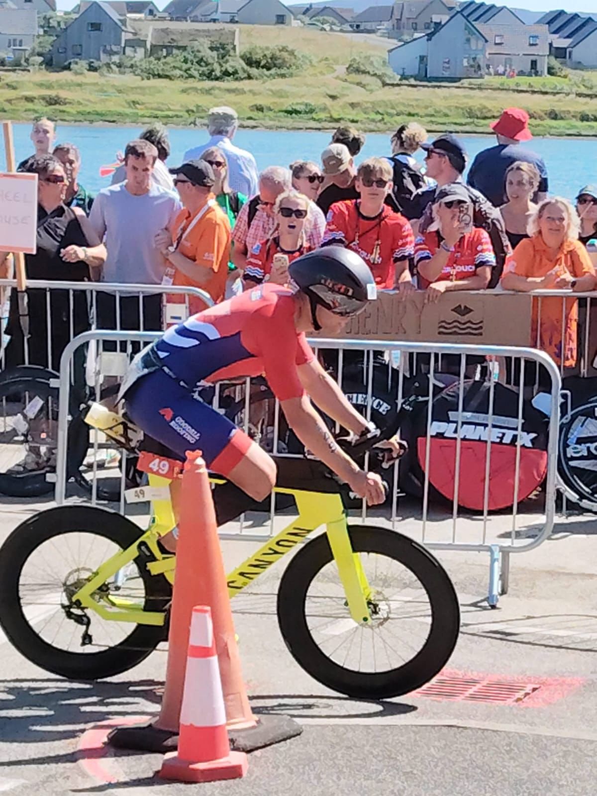 A cyclist wearing a helmet and racing outfit is riding a yellow Cannondale mountain bike during a race, with a crowd of spectators behind a metal barricade and a scenic background of water, green landscape, and houses.