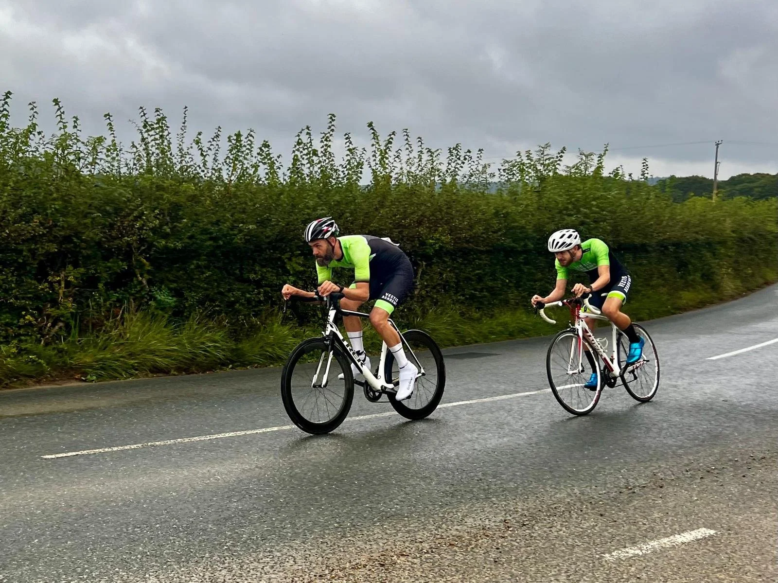 Two male cyclists riding racing bikes on a wet road with green bushes and overcast sky in the background.