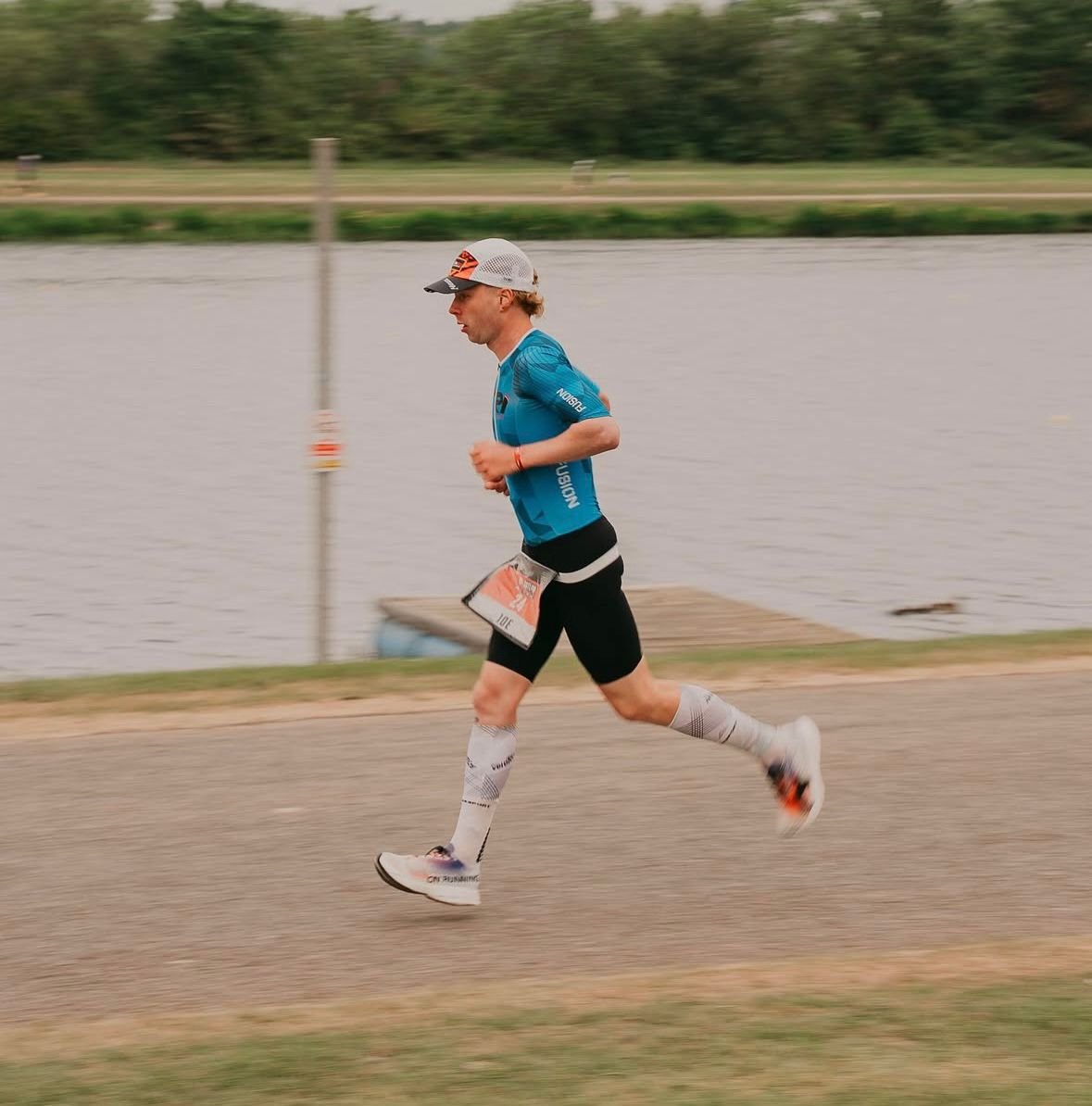 A man running on a paved path near a body of water, wearing athletic gear including a blue shirt, black shorts, a cap, and compression socks, during a race or outdoor event.