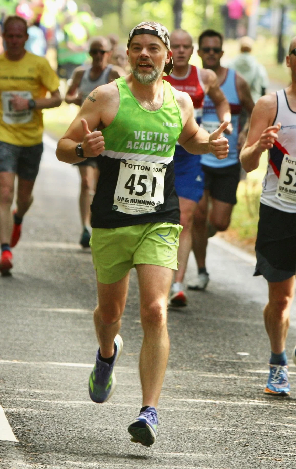 Man running in a marathon wearing a neon green tank top, green shorts, and a cap, with a bib number 451, giving a thumbs-up gesture.