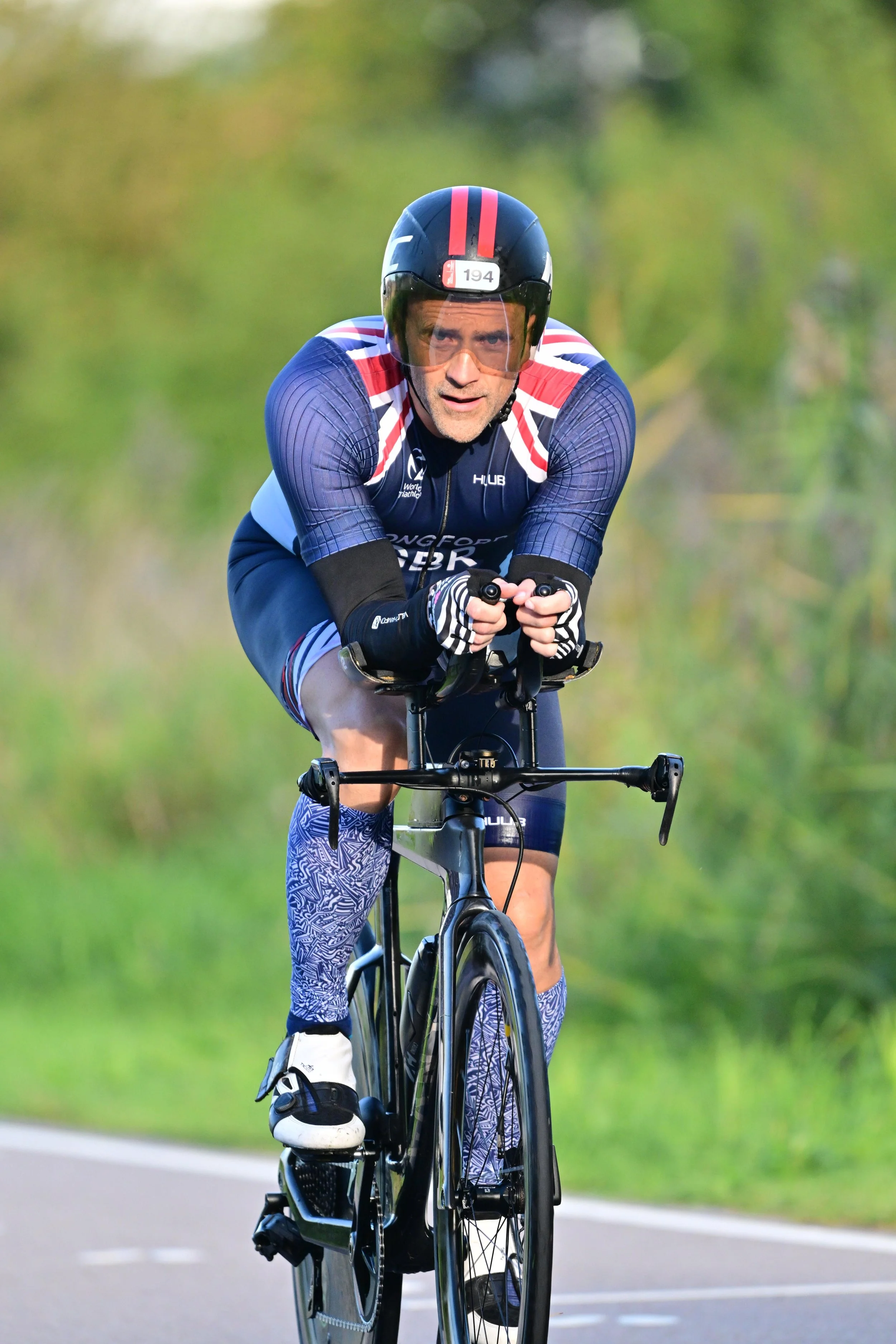 A man wearing cycling gear, including a helmet, sunglasses, gloves, and a jersey, rides a black road bicycle on a paved road with greenery in the background.