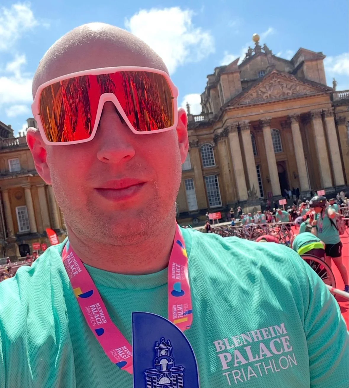 Man taking a selfie in front of Blenheim Palace during a triathlon event, wearing sports gear, sunglasses, and a medal.