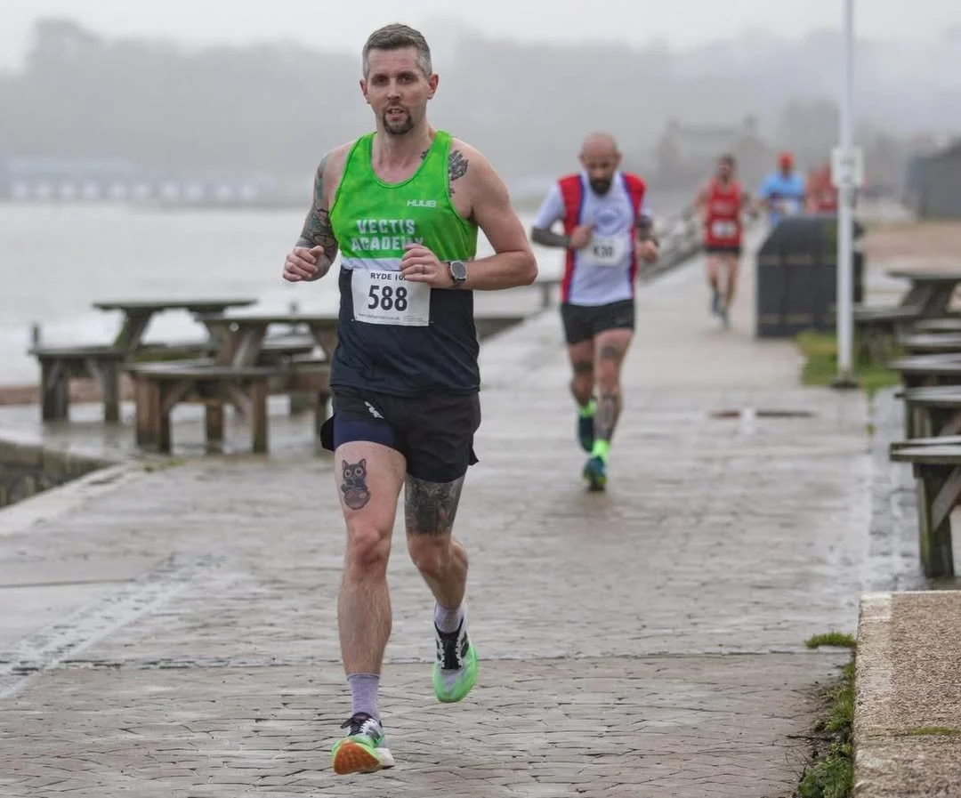 A man running in a race outdoors on a foggy day, wearing a green and black athletic tank top, black shorts, and neon green running shoes, with tattoos on his arms and legs.