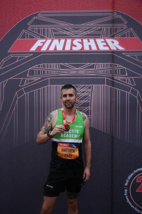 Male marathon runner standing in front of a finish line banner, holding his medal, smiling with tattoos on arms, wearing a green and black athletic outfit, with a bib number 2543.