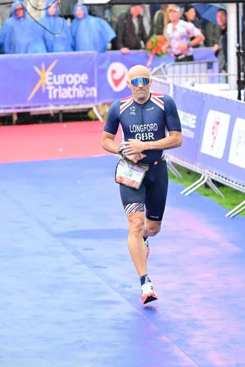 Male triathlete running on a blue and purple finish line at a triathlon event, wearing sunglasses, a blue uniform, and a race number bib.