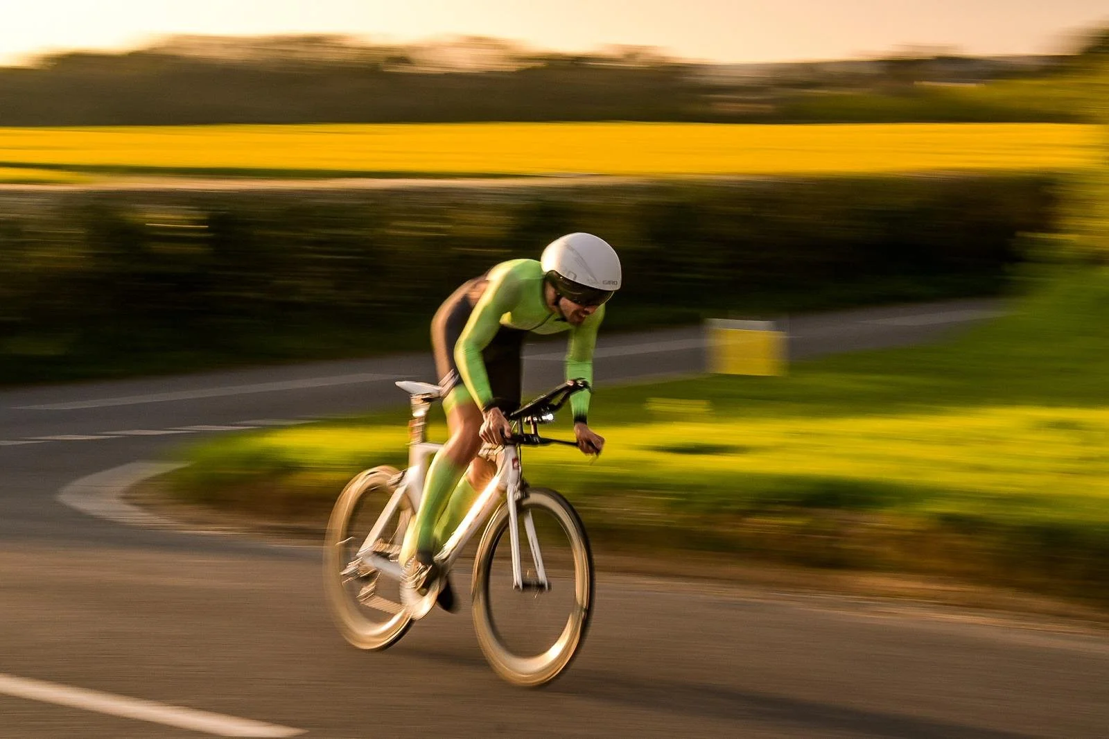 A cyclist wearing a green and black suit and a white helmet riding a bicycle on a paved road during sunset.
