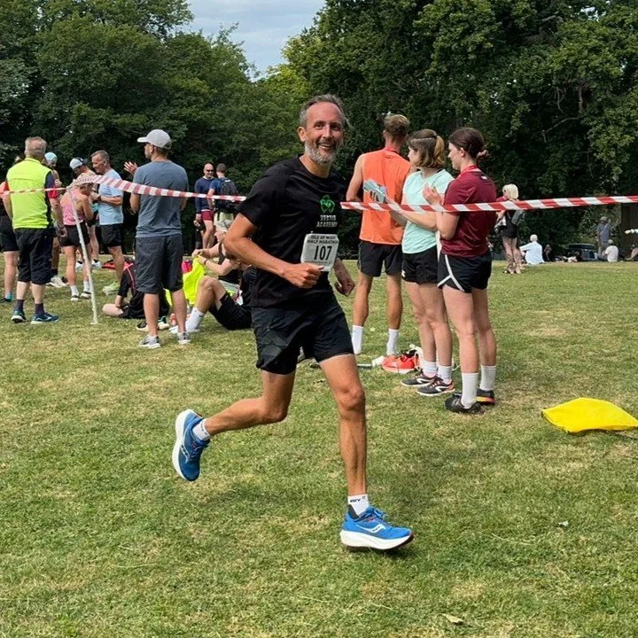 Man running in a race, wearing a black shirt and shorts, with a race bib number 107, smiling and looking at the camera. In the background, spectators and volunteers are gathered, some standing and some sitting on the grass, with trees in the distance