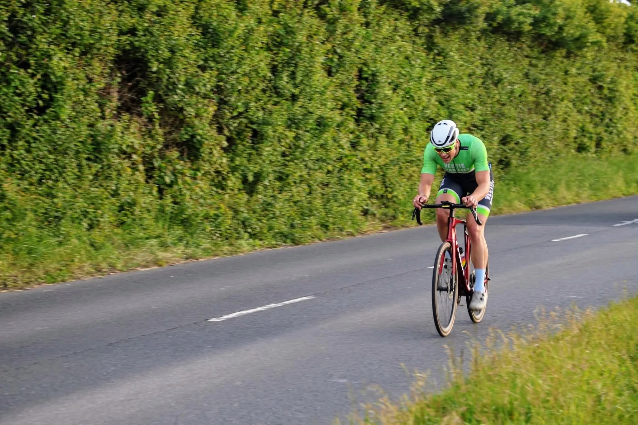 A cyclist in a green uniform with a white helmet riding a red bicycle on a paved outdoor road with green bushes on the side.