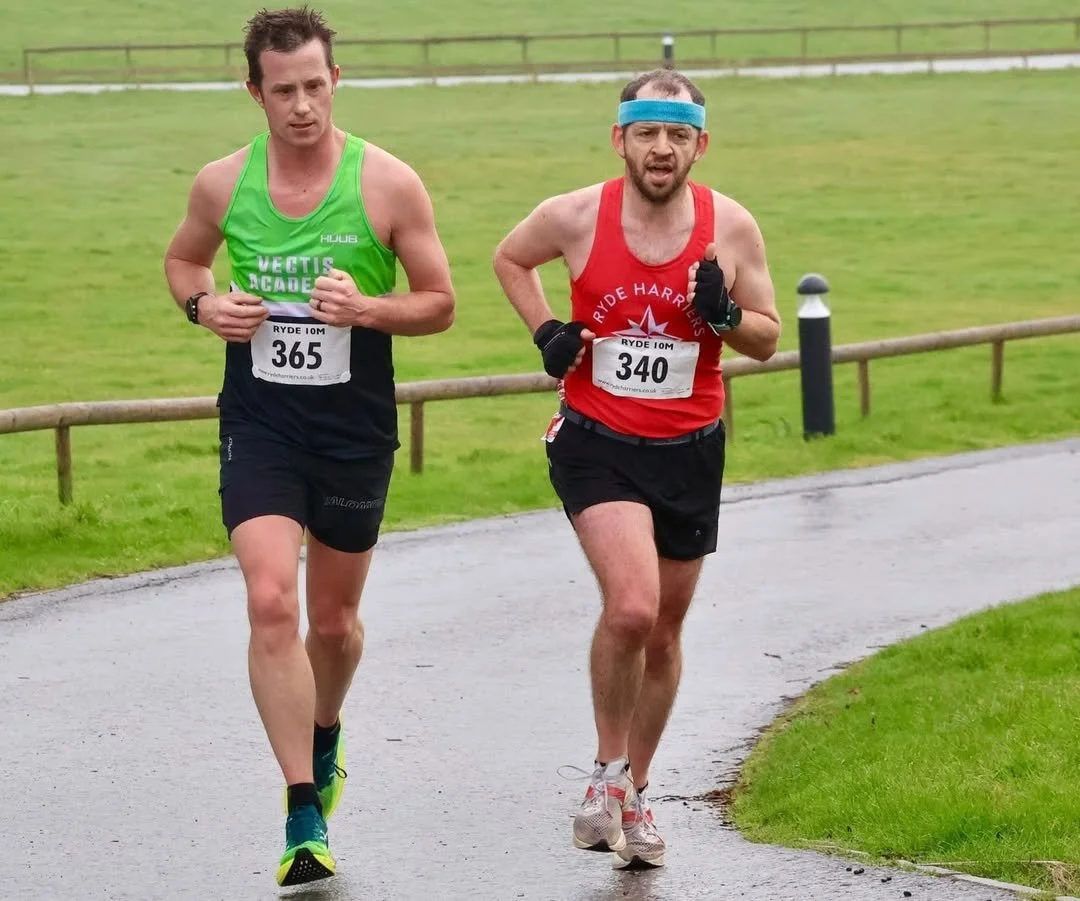 Two male runners participating in a race on a cloudy day, wearing race bibs, with green grass and a fence in the background.