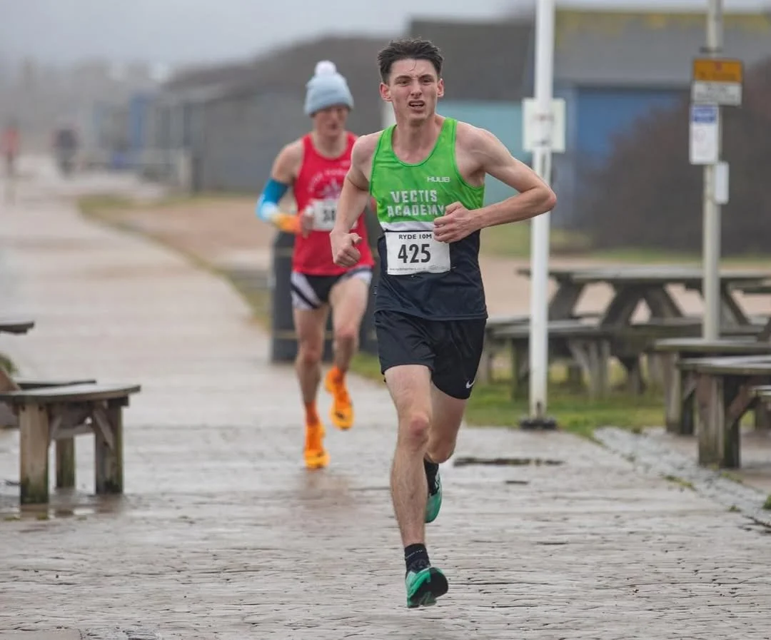A male runner with a green vest and black shorts competing in a race on a paved pathway, with a female runner in a red shirt and black shorts behind him, both outdoors on a cloudy day.