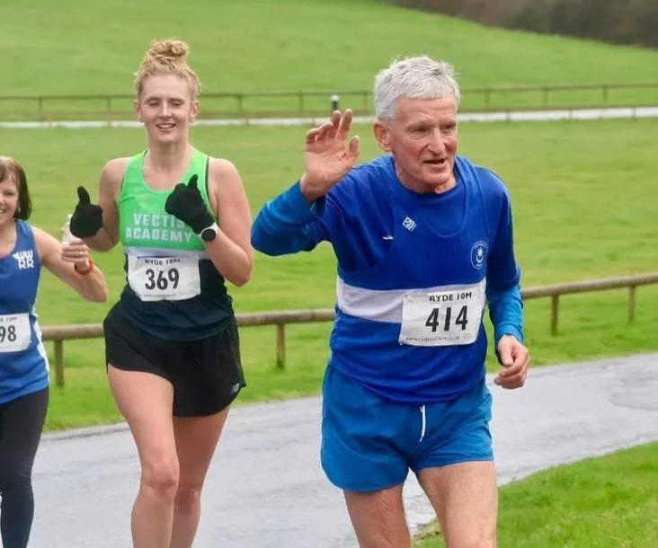 Older man running in a race, wearing a blue athletic shirt and shorts, waving his hand. Behind him, a young woman with a high bun, wearing a green and black athletic outfit with the number 369, giving a thumbs-up. Other runners are visible in the bac