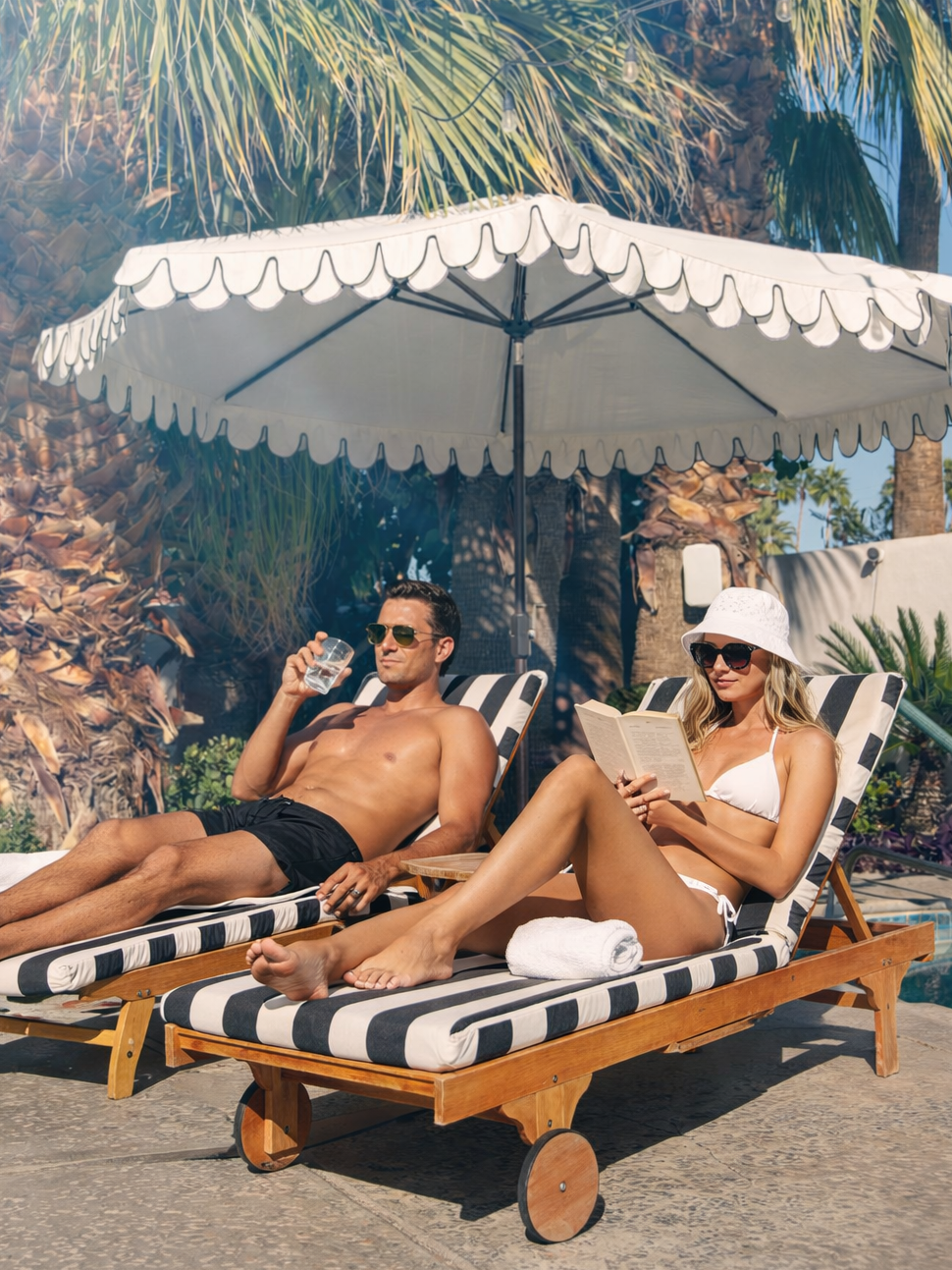 A man and a woman relaxing on lounge chairs beside a pool under a large parasol. The man is shirtless, wearing sunglasses and black swim trunks, holding a drink. The woman is in a white bikini, with a white hat and sunglasses, reading a book. Palm tr