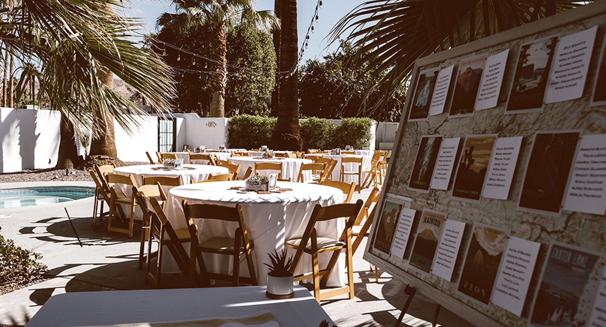 Outdoor event setup with round tables covered with white tablecloths, surrounded by wooden chairs, in a sunny patio area with palm trees and string lights. A menu board featuring various event posters is visible on the right.