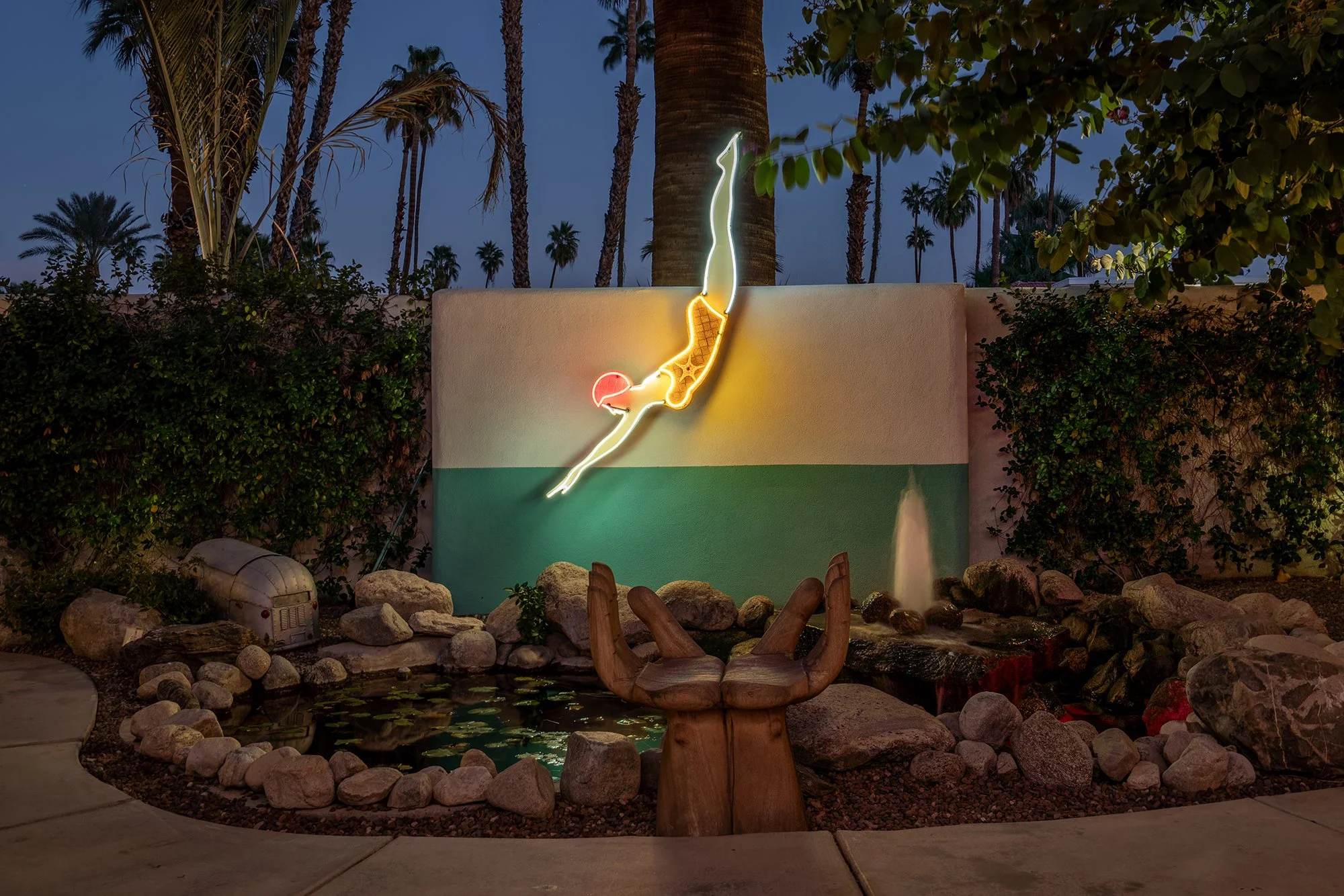Neon sign of a diver in mid-dive mounted on a wall, with a water fountain below, surrounded by rocks and a wooden hand sculpture, in a landscaped outdoor area with palm trees and bushes at dusk.